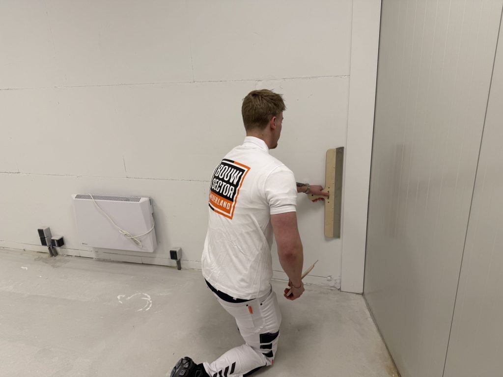 Geknielde stukadoor trekt met een brede spaan het onderste deel van een wand vlak, naast een elektrische kachel