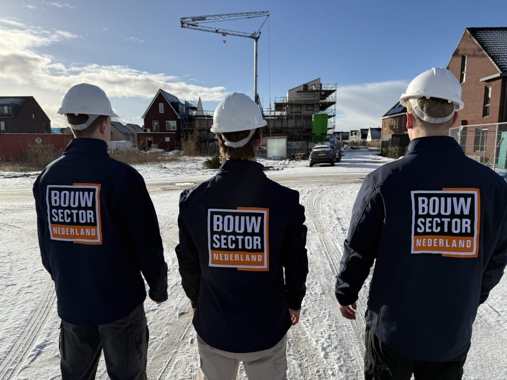Drie vakmensen van Bouwsector Nederland met witte helm kijken uit over een straat met woningen in aanbouw